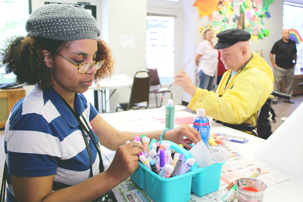 Structured Day Program, A woman with curly hair and glasses organizes markers at a table, while a man in a yellow jacket paints nearby.