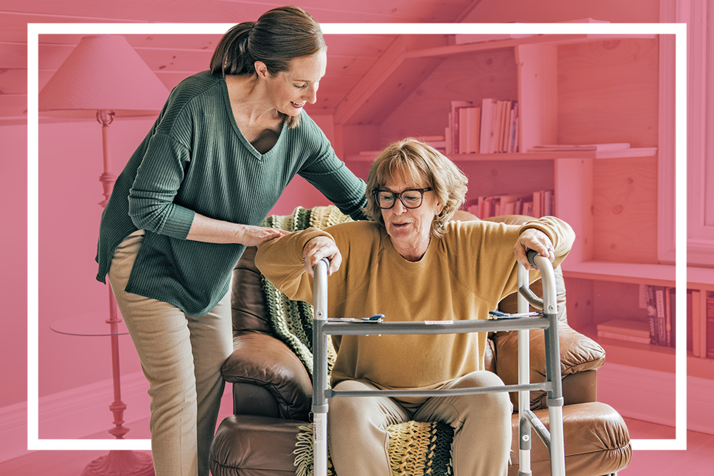 Middle-aged woman helps senior woman out of a chair with the aid of a walker.